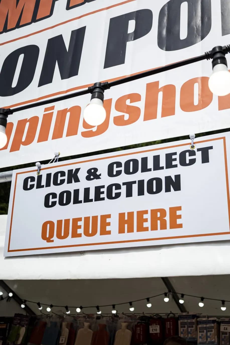 The click and collect sign hanging from the awning, reading Queue Here, with festoon lights strung across the front of the stall.