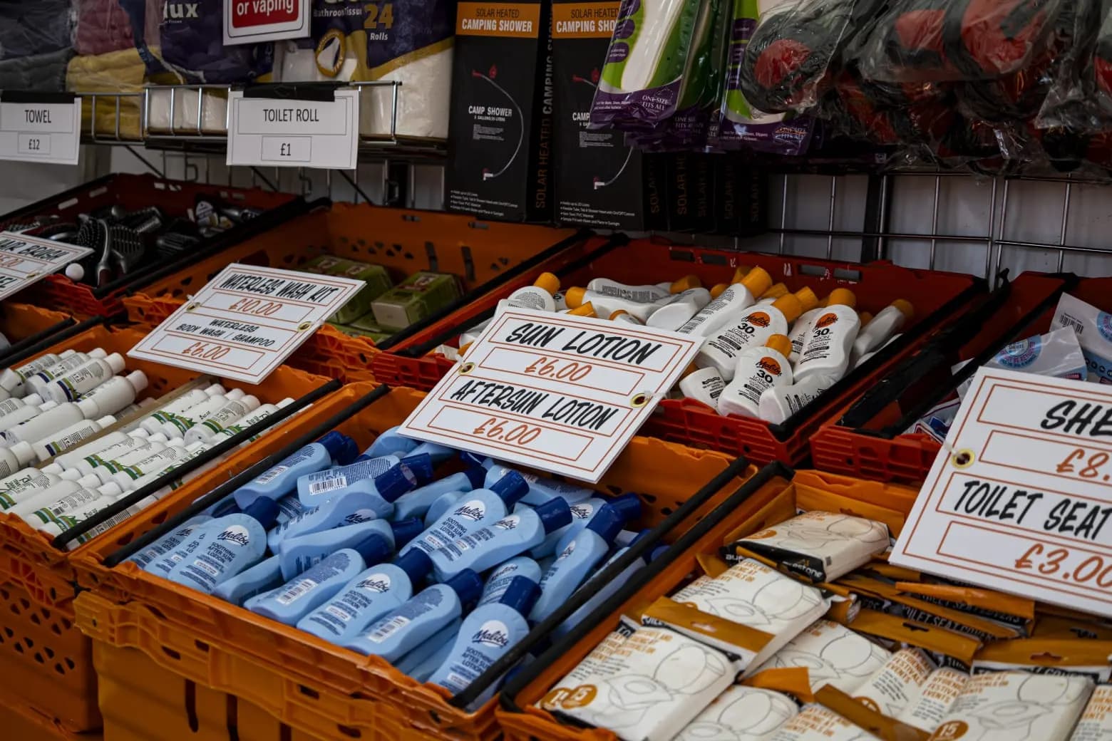 Crates of festival toiletries: sun lotion and aftersun, a waterless wash kit, toilet roll, towels and toilet seat covers, each with a handwritten price card.