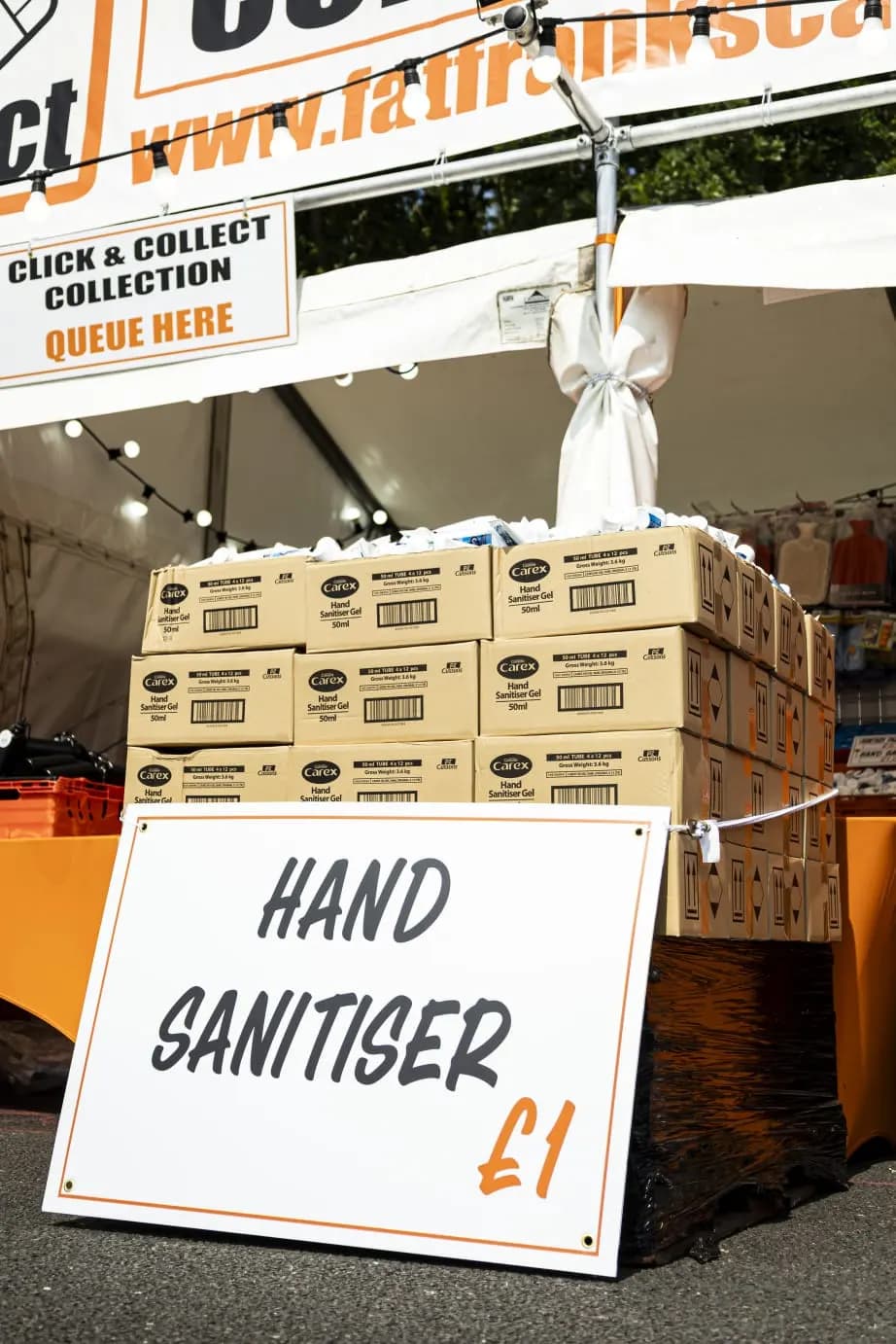 Boxes of hand sanitiser stacked behind a hand sanitiser price sign, under the click and collect sign.